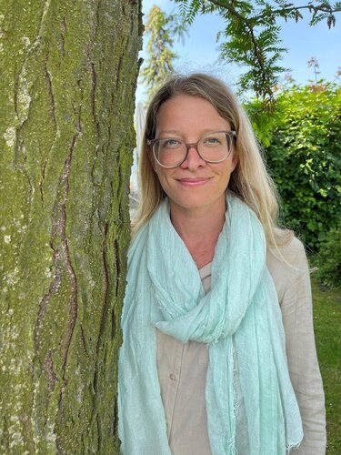 Frau mit Brille und Schal lehnt an Baum.