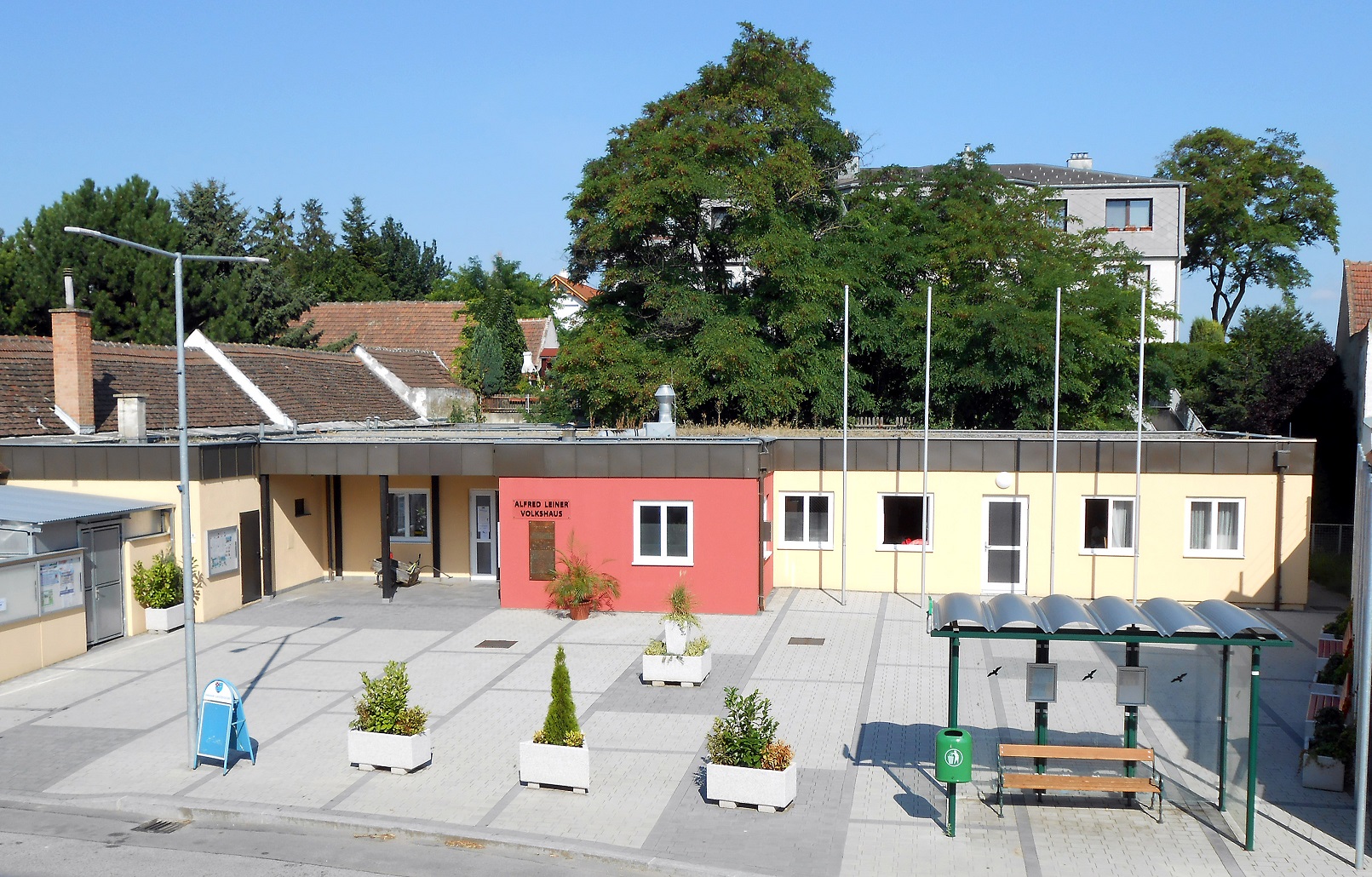 Rechts im Bild ist eine Bushaltestelle, daneben drei kleine Pflanzen und dahinter das Volkshaus in rot und beige.